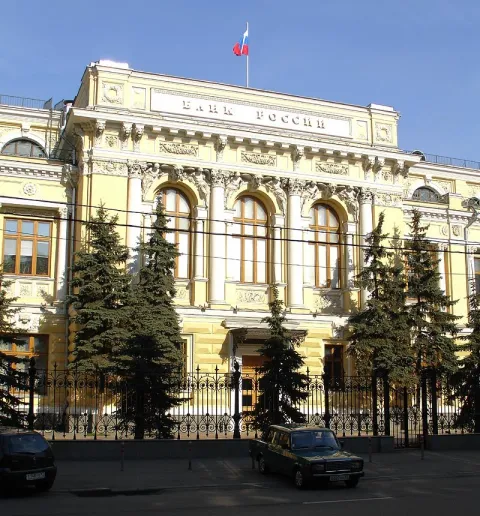 Фотография: светло-жёлтое здание с колоннами и надписью «Банк России» на фасаде, над ним российский флаг, перед зданием ели и ограда