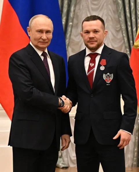 Фотография: Пётр Ян в тёмном костюме с наградой на лацкане пожимает руку Владимиру Путину на фоне флага