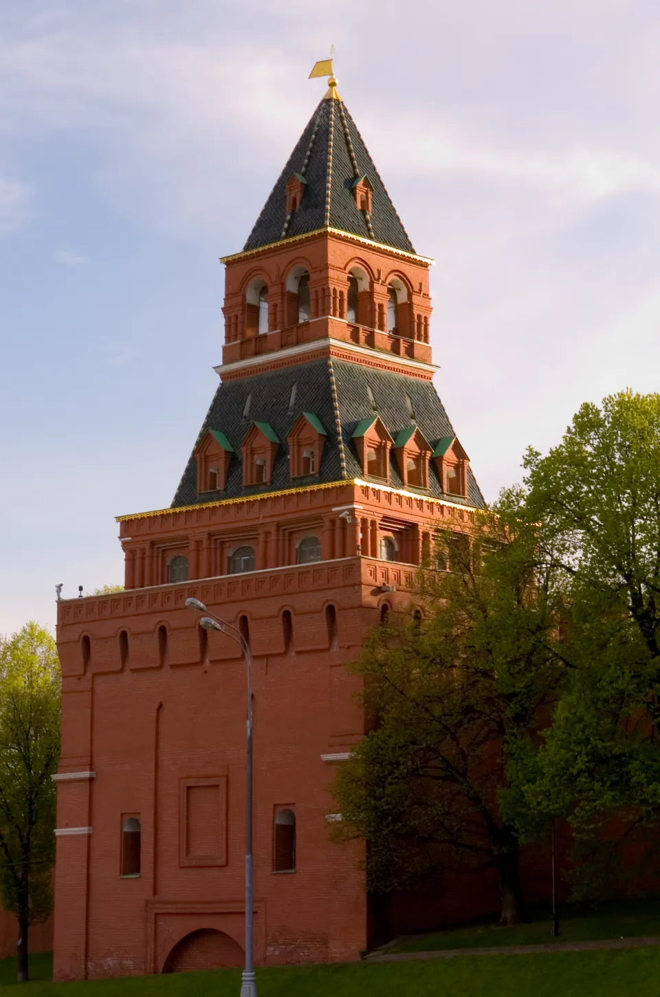 Краснокирпичная башня Московского Кремля с двухъярусным шатровым верхом и флюгером, рядом зелёные деревья
